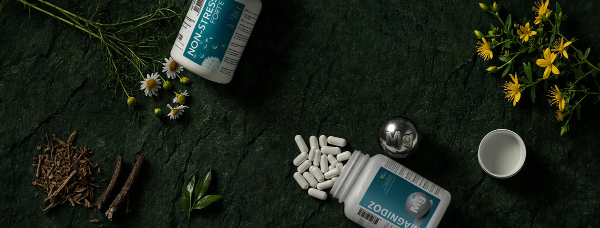 Two white supplement bottles labeled Magnidoz and Non-Stress Forte are arranged on a dark textured surface with herbs, flowers, and a small white cup. 